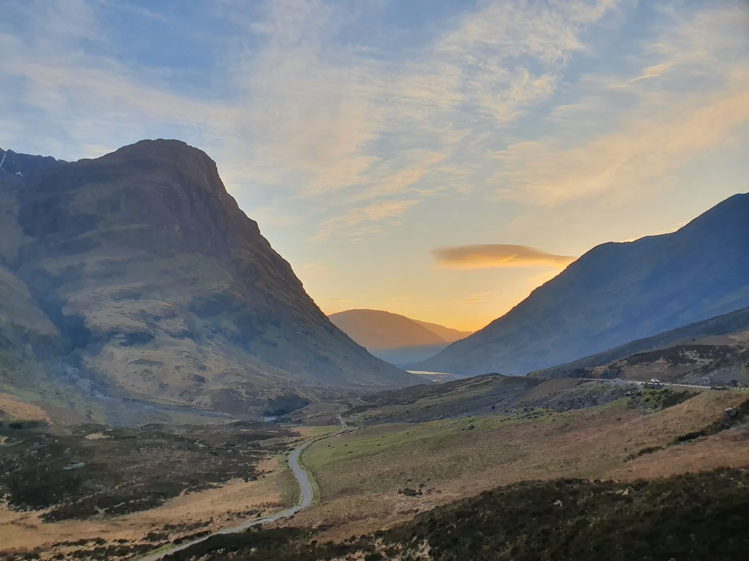 Sunrise light filling the Glencoe pass between Aonach Dubh and the Three Sisters, the old road winding along the valley floor towards a distant loch