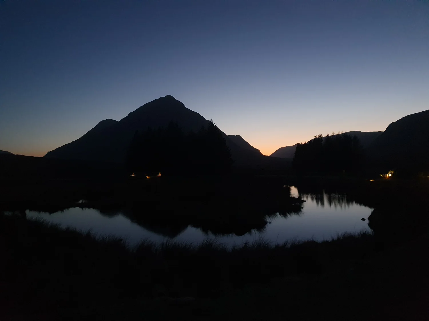 Buachaille Etive Mòr silhouetted at twilight, with the lit windows of Kingshouse and their reflection glowing on the lochan below