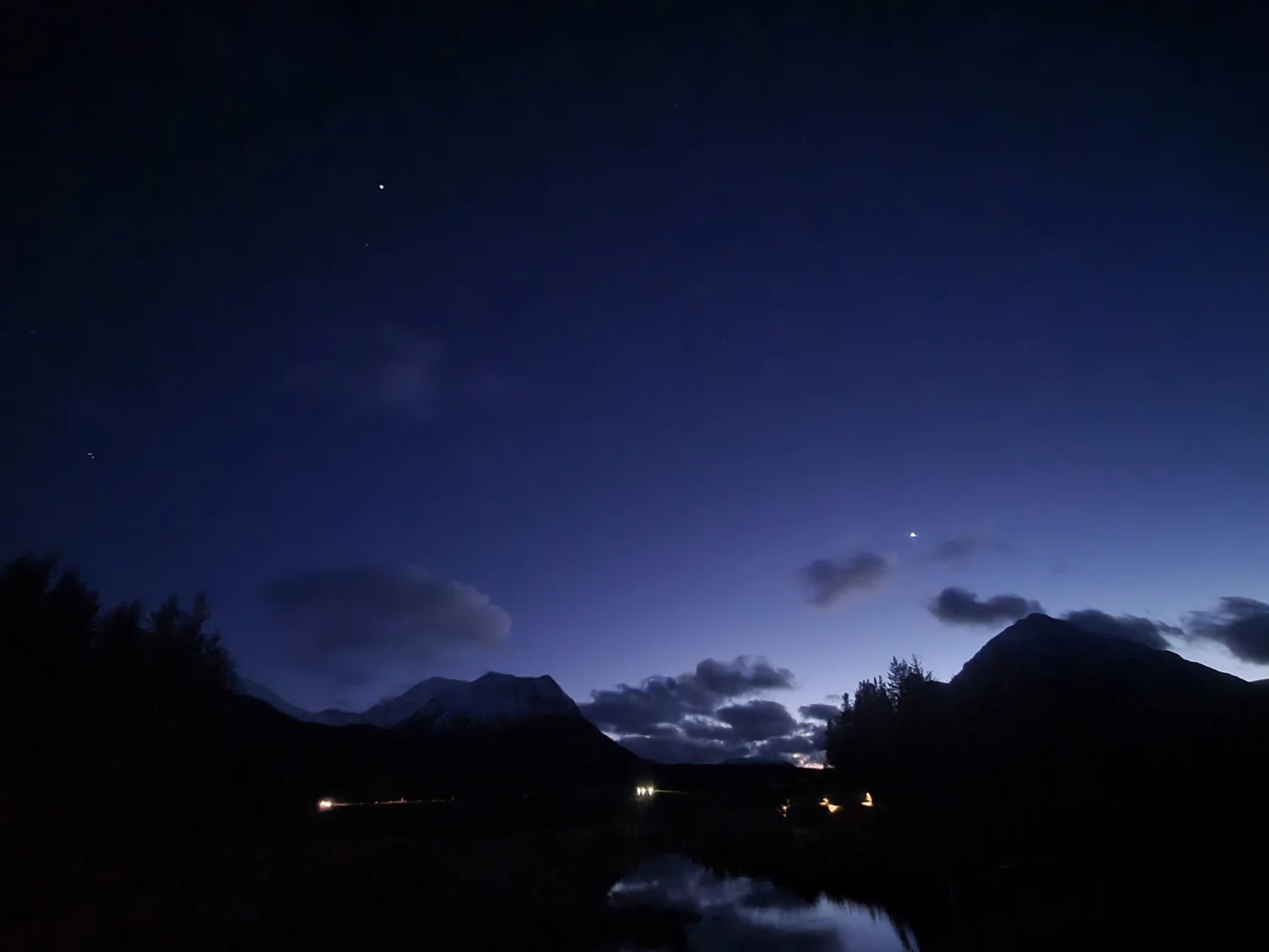 A planetary alignment visible in the twilight sky above the mountains, with cottage lights reflected in the still water below