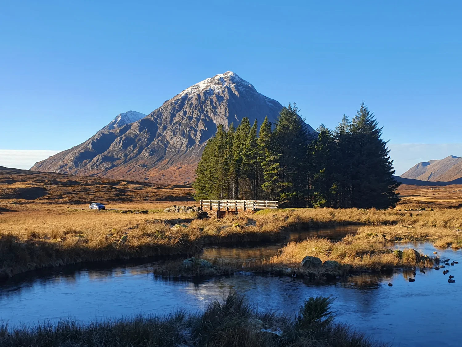 Snow-capped Buachaille Etive Mòr above the pines and footbridge at the Kingshouse lochan, with a car parked on the moorland road below